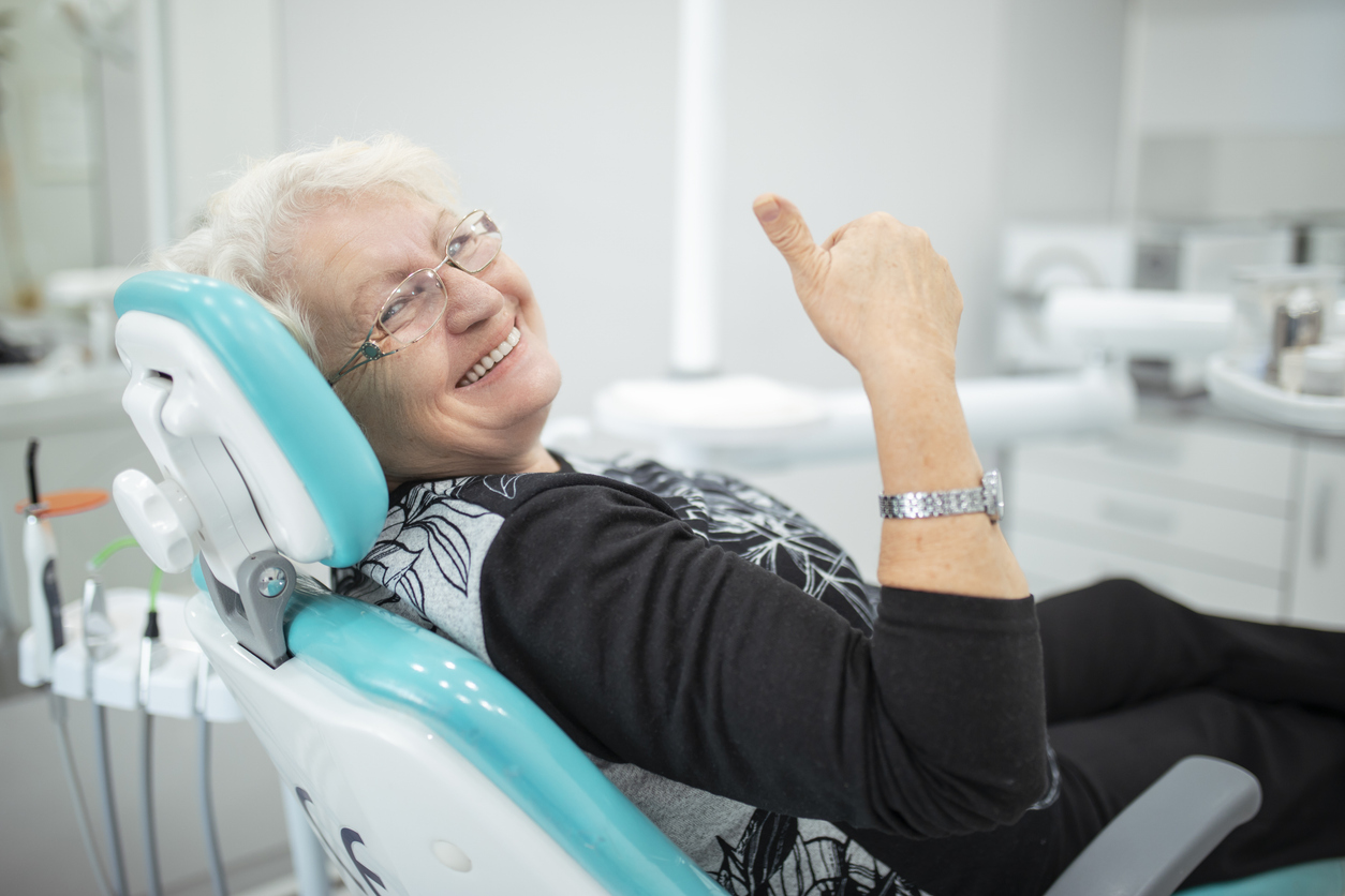 The image shows a senior woman sitting in a dental chair with a thumbs up to represent if dental implants are permanent.